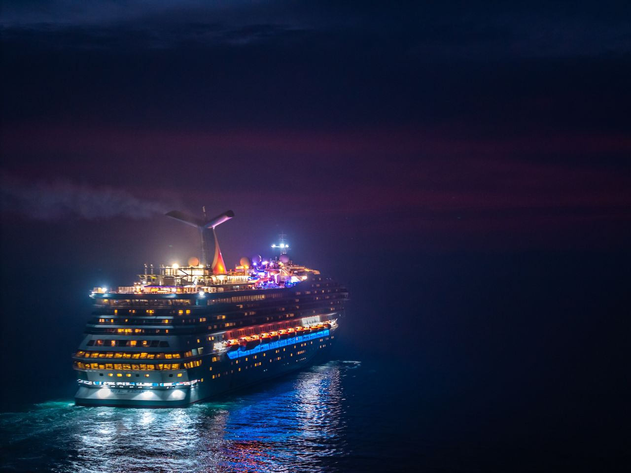 services-03 A brightly lit cruise ship sails through the ocean under a vivid night sky.